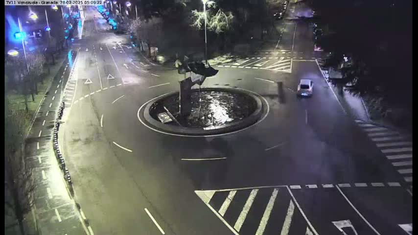 A nighttime aerial view of a roundabout with a single car on the road, surrounded by dark trees and streetlights.