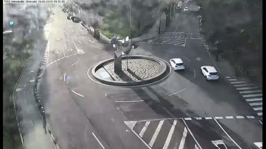 A high-angle view of a roundabout with two white cars, surrounded by roads and trees on an overcast day.