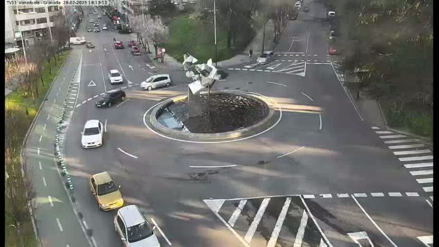 A roundabout with several cars driving around it, surrounded by roads and greenery under a sunny sky.