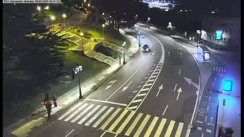 A nighttime street scene shows a curved road with crosswalks, two pedestrians, and two vehicles, with steps and greenery alongside the road, all lit by streetlights.
