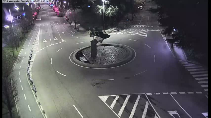 An aerial nighttime view shows a roundabout with a sculpture in the center, surrounded by roadways and pedestrian crossings.