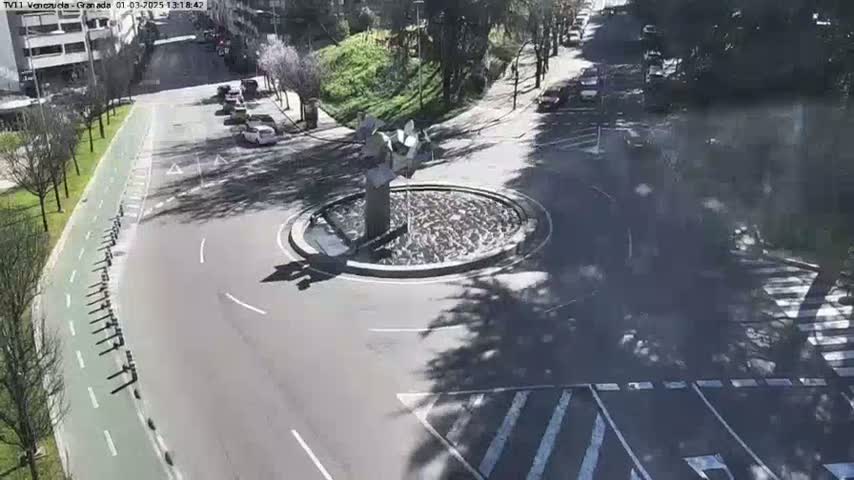 A sunny day reveals a traffic circle with a modern sculpture in the center, surrounded by a road with parked cars and a bike lane next to a grassy area with trees.