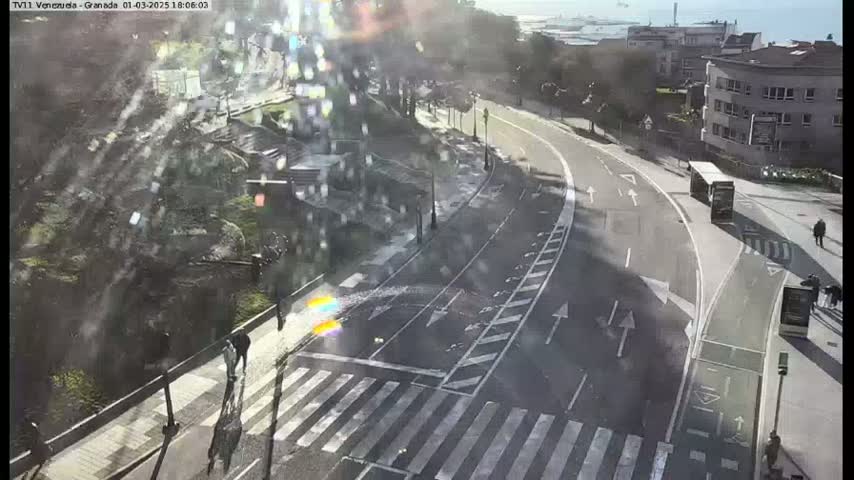 A sunny day overlooking a curved road with crosswalks, pedestrians, and a bus, situated near buildings and greenery.