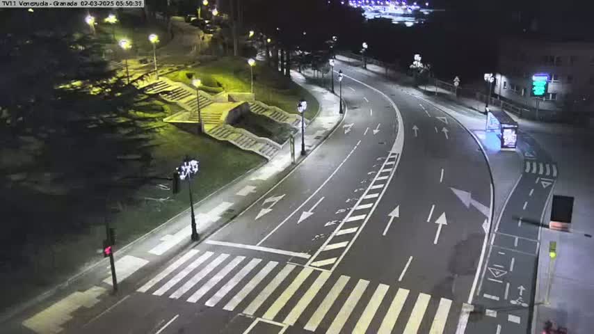 A nighttime, high-angle view shows a curving road with pedestrian crosswalks, streetlights, and stairs leading up a grassy hill to the left.
