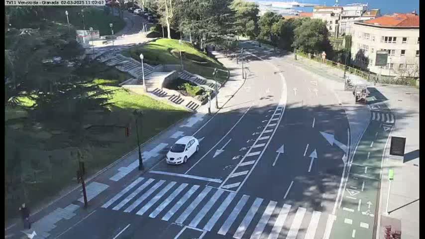 A sunny day shows a white car driving through a city intersection with pedestrian crosswalks and bike lanes, alongside green spaces and buildings.