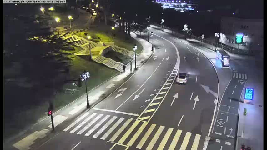 A nighttime, well-lit roadway curves around a pedestrian area with stairs and a few people walking, near some buildings.