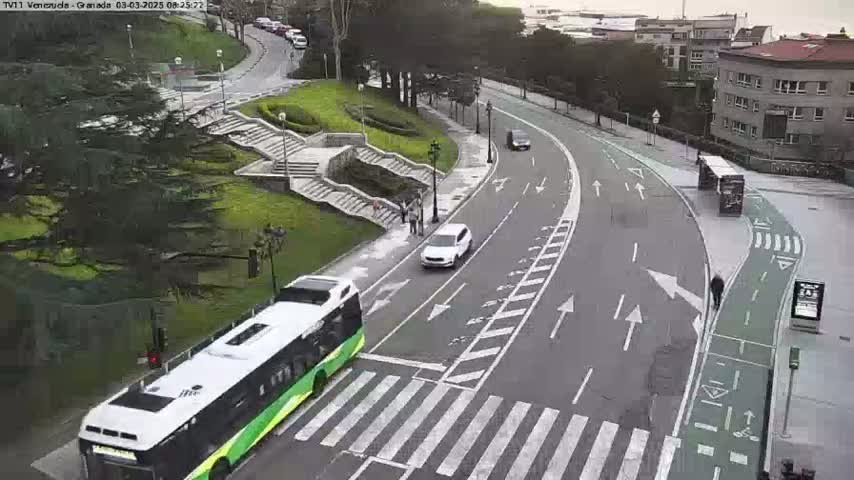 A green and white bus is parked near a crosswalk on a curved road next to a stairway and a grassy area with some people walking; a white car is also visible on the road, under mostly cloudy skies.