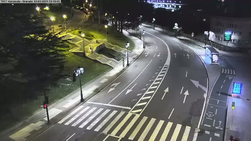 A nighttime, illuminated view of a curved roadway with crosswalks, stairs leading down to a grassy area with trees, and a building in the background.
