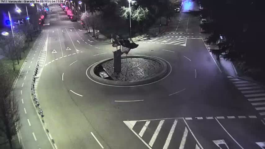 A nighttime aerial view shows a roundabout with a dark sculpture in the center, surrounded by roads with minimal traffic.