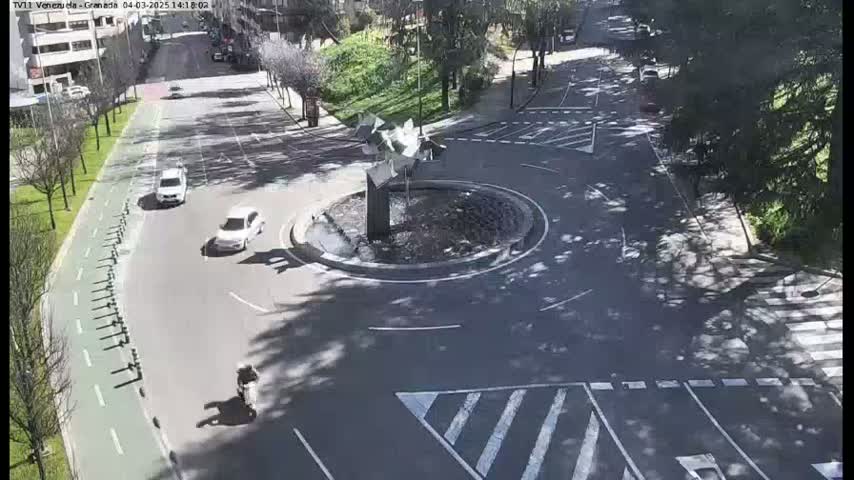 A sunny day shows a circular traffic island with a modern fountain in the center of a roadway intersection, with a few cars and a motorcyclist visible.