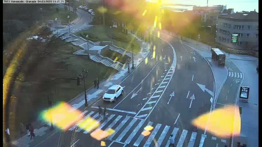 A sunny, outdoor scene shows a curved road with pedestrians crossing at a crosswalk, a white SUV, a bus, and a staircase leading to a grassy area.