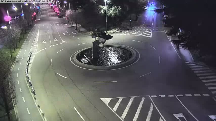 A nighttime aerial view of a roundabout with a dark sculpture in the center, surrounded by roadways and pedestrian crosswalks.