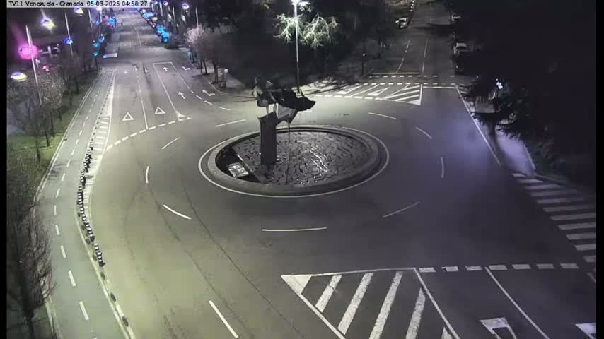An aerial nighttime view of a roundabout with a sculpture in the center, surrounded by roads and pedestrian crossings.