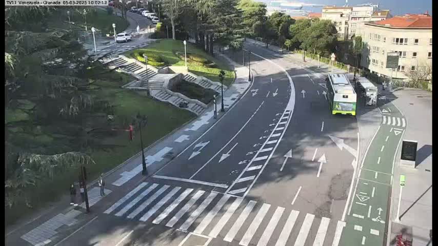 A paved road with crosswalks curves through a park-like area with stairs and greenery, under a sunny sky; a bus is stopped near a building.