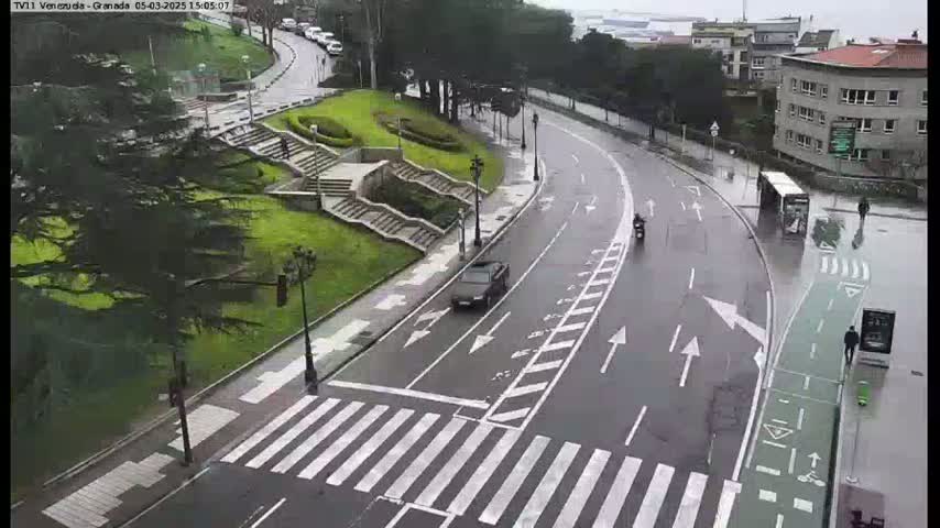 A wet road curves through a city scene, passing by a pedestrian crossing, a grassy area with stairs, and several buildings, with a few vehicles and pedestrians visible.