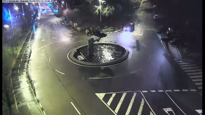 A nighttime, wet, overhead view of a roundabout with a fountain in the center, surrounded by roads and some parked cars.