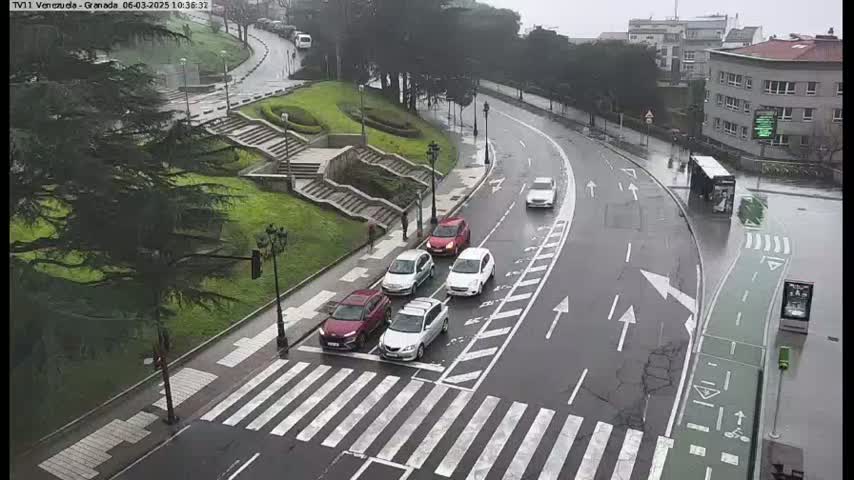 Several cars are driving on a wet road that curves around a grassy area with stairs, under a gray sky.