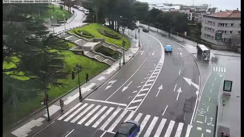 A wet road curves through a city scene, with pedestrians crossing a crosswalk and a few vehicles driving on the road, alongside a grassy area with stairs and trees.