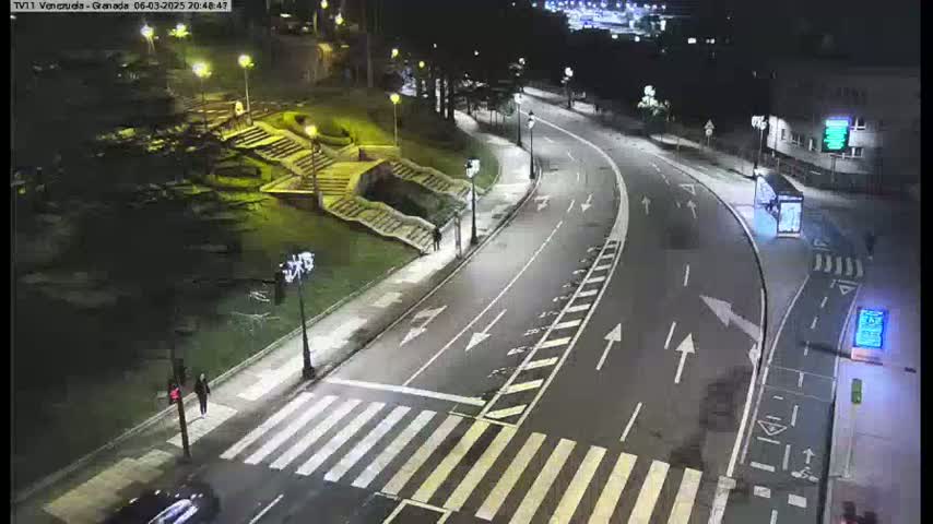 A nighttime street scene shows a curving road with a pedestrian crosswalk, streetlights, and a few people walking near a staircase and green landscaping.