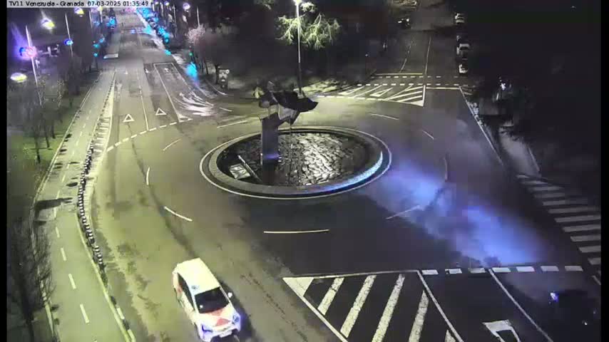 A nighttime aerial view shows a roundabout with a dark sculpture in the center, surrounded by streets with a few cars and pedestrian crossings.