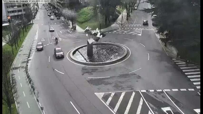 A roundabout with a modern sculpture in the center, surrounded by several cars and a cyclist on a grey, overcast day.