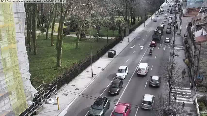 A city street scene on a cloudy day shows several cars driving in both directions along a roadway that runs alongside a park with many bare trees.