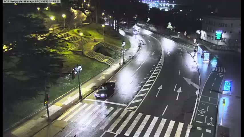 A dark car is stopped at a crosswalk on a wet, nighttime road curving through a park-like area with stairs and streetlights.