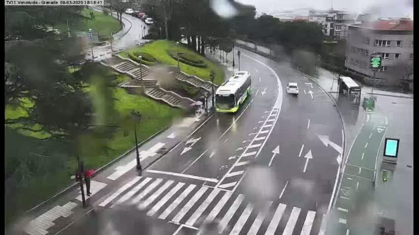 A rain-slicked road curves around a grassy area with stairs, featuring a bus and several cars, with a pedestrian crossing visible.