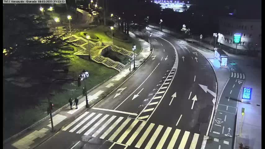 A nighttime, high-angle view of a roadway curving through a landscaped area with stairs, featuring a crosswalk and a few pedestrians.