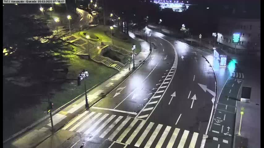 A wet, empty road curves through a nighttime cityscape, passing by pedestrian crossings and landscaped areas with stairs.