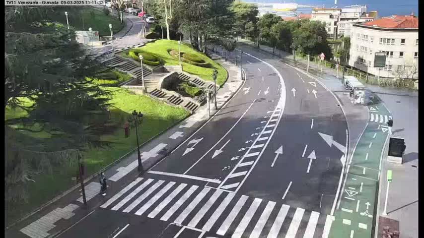 A sunny day shows a curved road with pedestrian crosswalks, lined with green space, stairs, and buildings in the background.