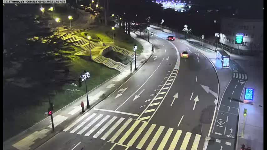 A nighttime view of a curved roadway with pedestrian crossings, stairs, and several cars, illuminated by streetlights.