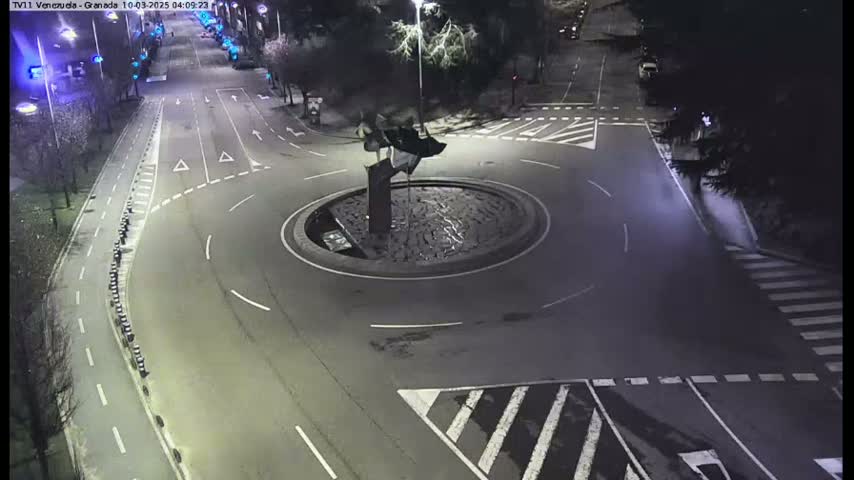 A nighttime aerial view of a traffic circle with a dark sculpture in the center, surrounded by roads and pedestrian crossings.