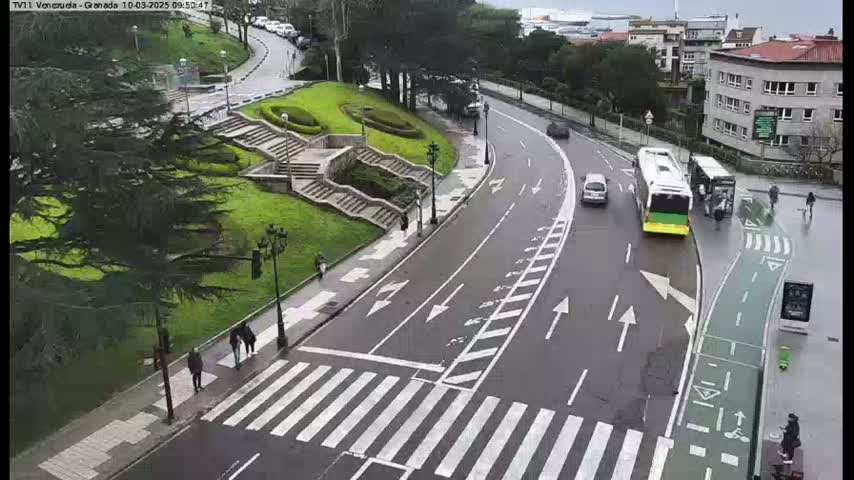 A wet road curves through a city scene, featuring a bus stop, a crosswalk, and a bike lane next to a grassy area with stairs.