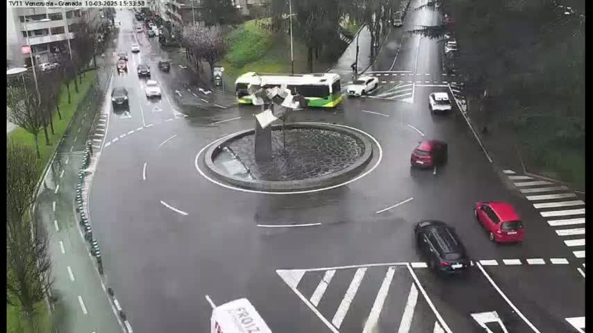 A wet roundabout with several cars and a bus, surrounded by city streets and buildings on a rainy day.