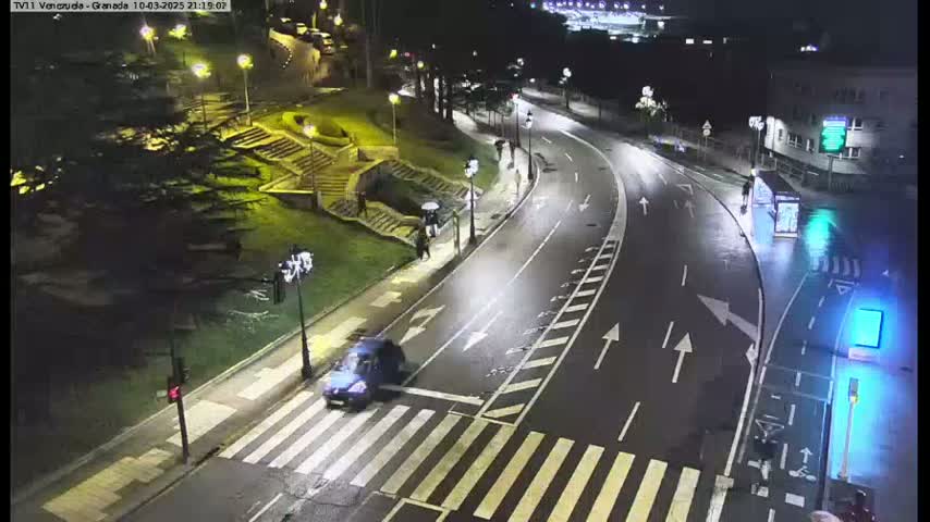 A wet, nighttime street scene shows a curving road with pedestrian crosswalks, a few people walking, and a dark-colored car crossing an intersection.