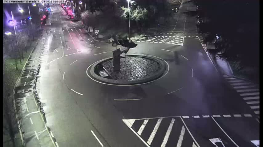 A wet roundabout at night, featuring a dark sculpture in the center, is surrounded by roads with minimal traffic.