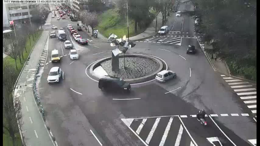 A roundabout with several cars and a motorcycle, situated in a city with wet roads.
