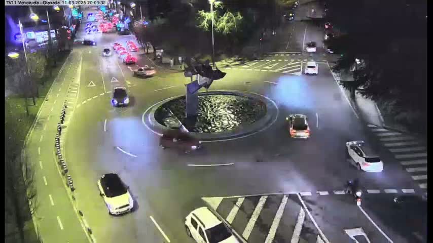 At night, several cars drive around a traffic circle featuring a dark fountain, with a pedestrian and a motorcycle also visible.