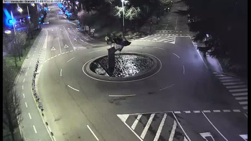 A nighttime aerial view shows a roundabout with a water feature and sculpture in the center, surrounded by roads and pedestrian crossings.