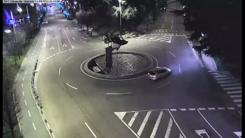 A car drives through a roundabout at night, illuminated by streetlights, with a dark sculpture in the center.