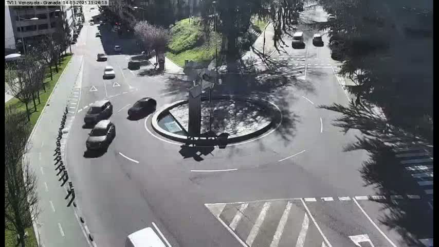 Several cars are navigating a roundabout in a sunny, urban setting surrounded by trees.