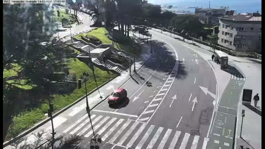 A sunny day shows a curving road with a bike lane, pedestrian crosswalks, a red car, and stairs leading down to a grassy area with trees and buildings in the background.
