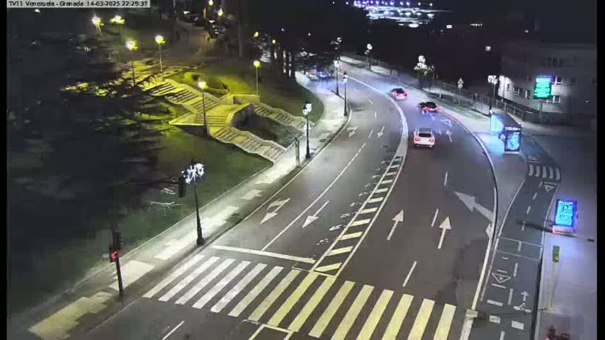 A nighttime, well-lit street curves past a set of stairs and park-like area, with a few cars visible on the road.