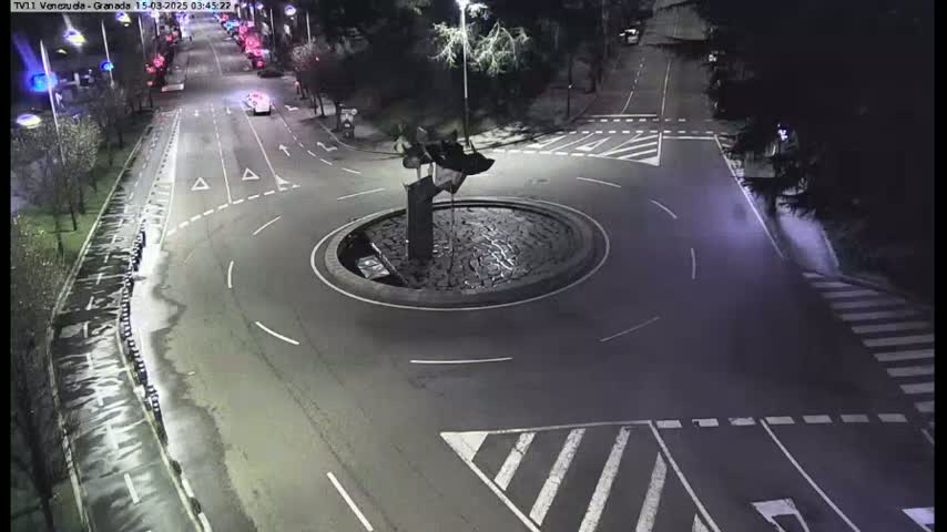 A nighttime overhead view shows a roundabout with a dark, abstract sculpture in the center, surrounded by wet roads and crosswalks.