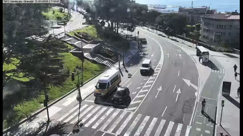 A sunny day reveals a curving road with several vehicles, including a van and cars, near a pedestrian crossing and stairway, surrounded by green landscaping and buildings.