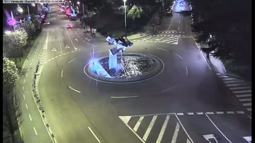 A nighttime aerial view shows a roundabout with a modern sculpture in the center, surrounded by streets and crosswalks.