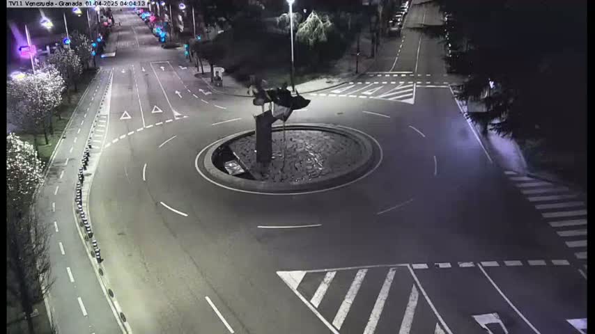 An aerial nighttime view shows a traffic circle with a dark sculpture in the center, surrounded by roads with minimal traffic.