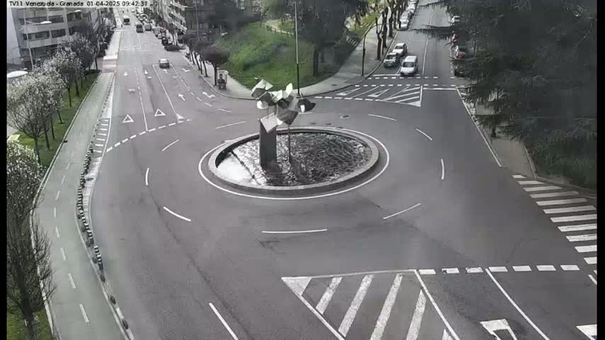 A traffic roundabout with a modern sculpture in the center, surrounded by roads with some cars and pedestrian crossings, on a partly cloudy day.