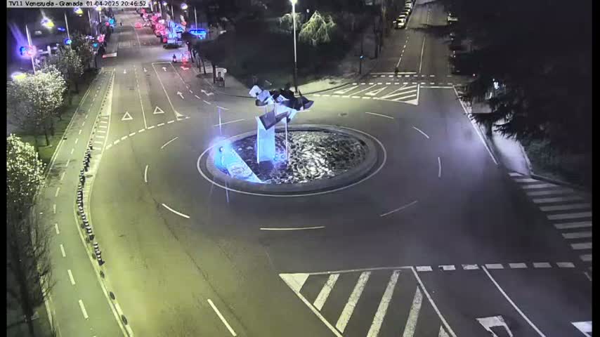 A nighttime aerial view shows a roundabout with a modern sculpture in the center, surrounded by roads with minimal traffic.